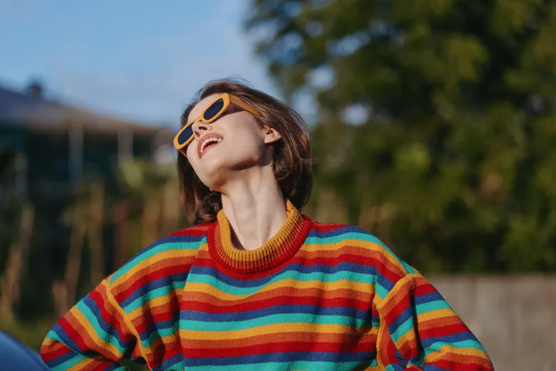 Young woman wearing a colorful striped sweater and yellow sunglasses, smiling and leaning back outdoors in a sunlit portrait, short brown hair and joyful expression for casual lifestyle fashion.