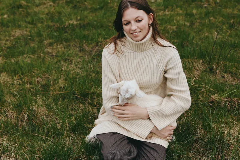 A woman in a cream sweater sits on a grassy field, gently holding a young goat kid. A lifestyle scene showing calm care, bonding, and rural simplicity in bright daylight.