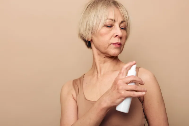 Mature woman with short blonde hair sprays deodorant on her underarm wearing a beige tank top against neutral background. Concept of personal hygiene, skincare, and self-care routine.
