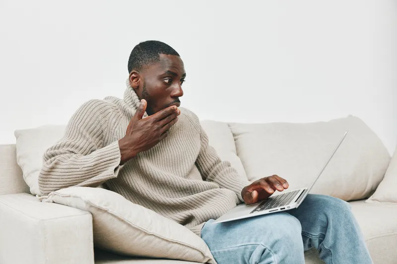 Black man in casual sweater looking shocked while working on a laptop, reflecting surprise and engagement in a modern home setting