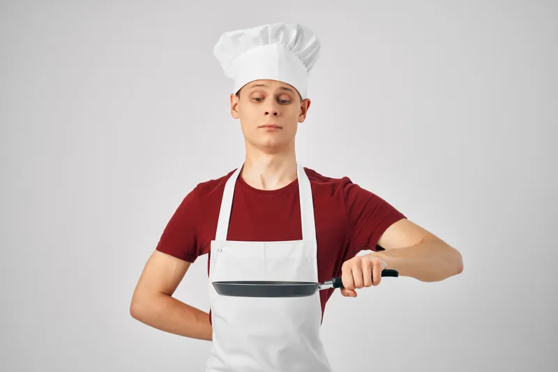 Мужчина в поварской форме держит сковороду в руках на профессиональной кухне, занимаясь приготовлением пищи