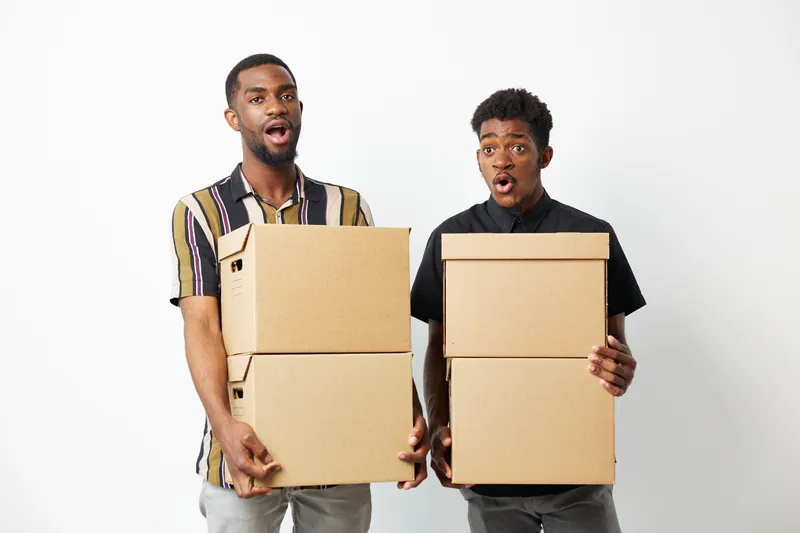 Two black men joyfully carrying cardboard boxes, showcasing excitement and teamwork while delivering goods. Perfect for diverse representation in commercial use.