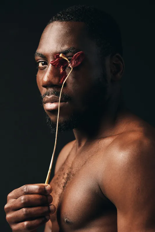 African man with dark skin holding dried flower near face in creative conceptual visual style. Portrait with artistic expression and bare chest on dark background for beauty and emotion.