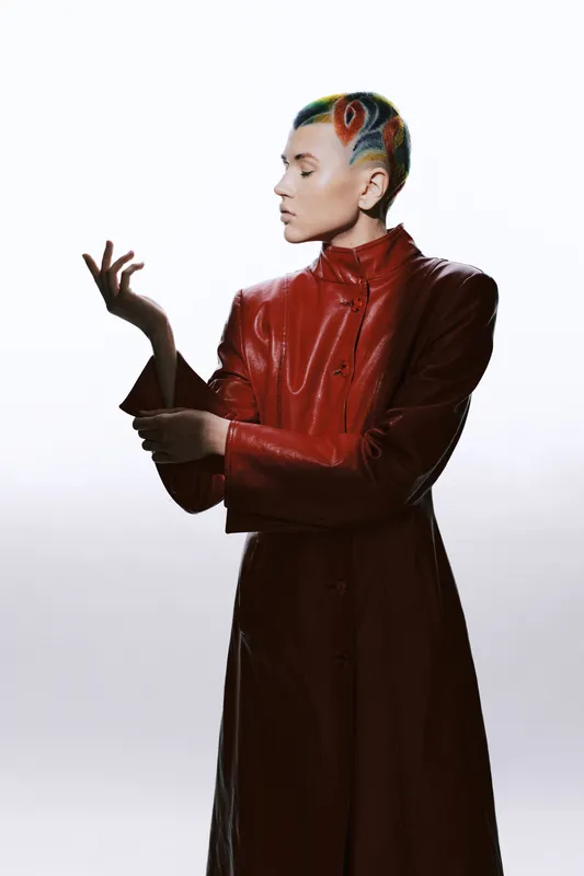 woman with colorful shaved hair wearing a red leather coat, standing in a side pose with white background, modern style, artistic fashion portrait