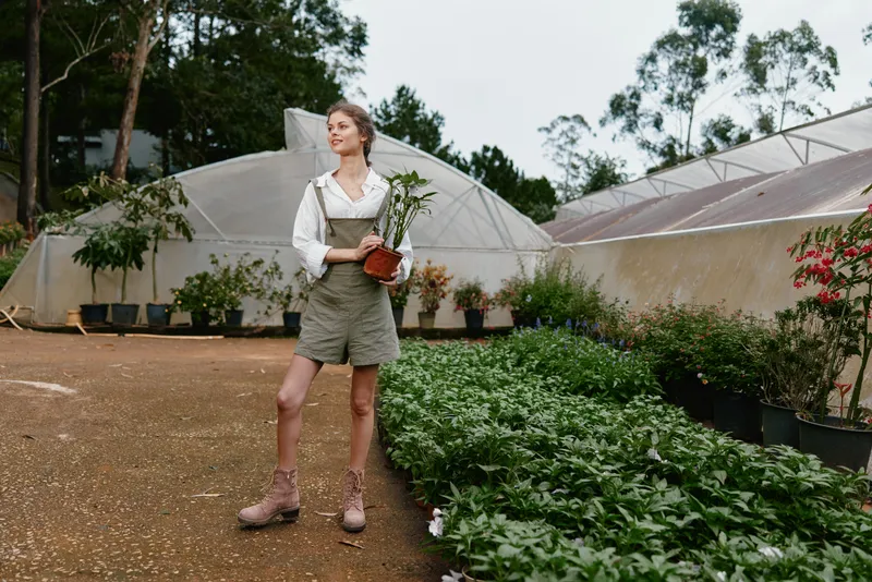Young woman in green overalls holding a potted plant in a greenhouse, surrounded by lush greenery, exuding a sense of tranquility and connection to nature