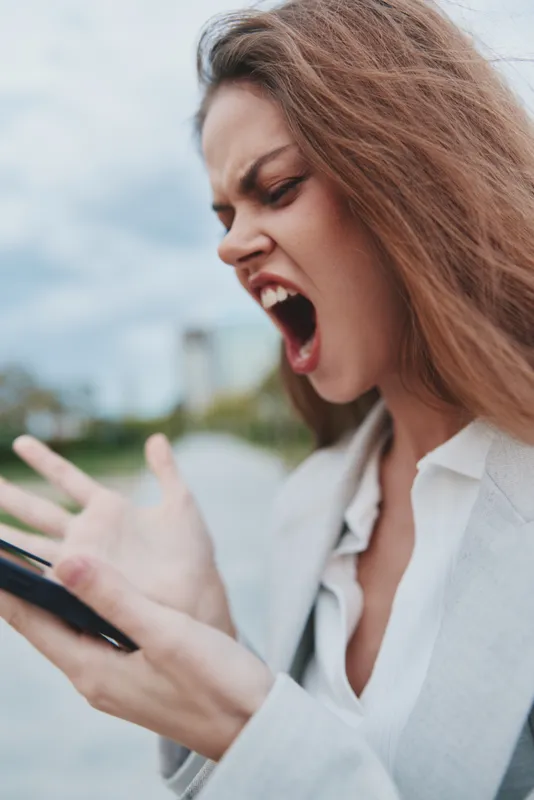 Upset businesswoman yelling at her smartphone outdoors, dressed in a light-colored blazer, showcasing frustration and determination in a city park.