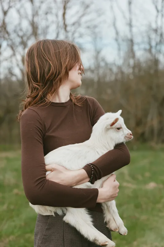Cheerful woman outdoors holding a white goat kid, warm sunlight in a green meadow, casual outfit, gentle bond with animal, natural colors, serene countryside vibe