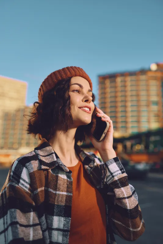 Woman on phone smiling in urban street at golden hour glow, candid lifestyle portrait with beanie and plaid jacket, conveying authenticity, mindful living and emotional storytelling vibe.