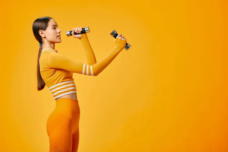 Woman fitness portrait in a vibrant yellow studio, wearing matching crop top and textured leggings, holding small dumbbells, demonstrating controlled strength training, focused posture and energy.