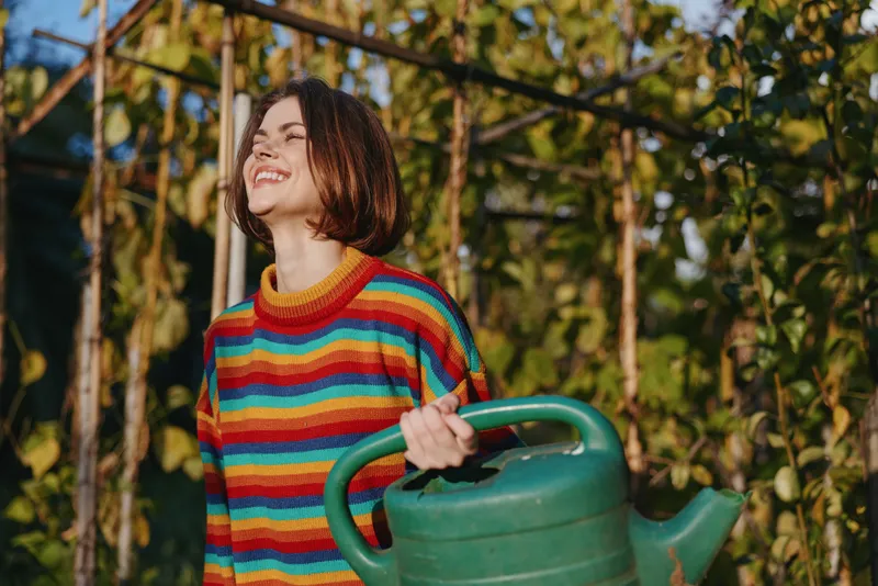 Young woman in a colorful striped sweater holding a green watering can in a sunlit garden, smiling and laughing with a short bob haircut, casual outfit for home gardening lifestyle.