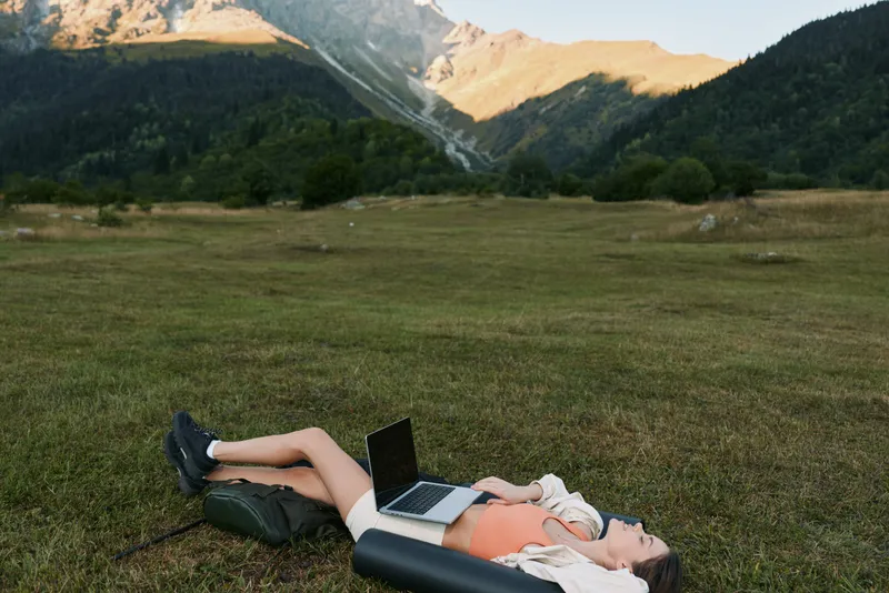 A person lies on a grassy field with a laptop open beside them, enjoying nature while working remotely. The scene conveys relaxation, focus, and productivity in an outdoor setting.