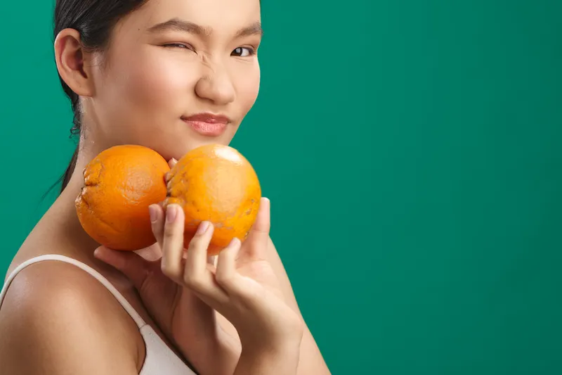 asian woman holds vibrant oranges in a bright studio against a green backdrop, smiling softly. the image conveys health, vitality, and a fresh lifestyle vibe.