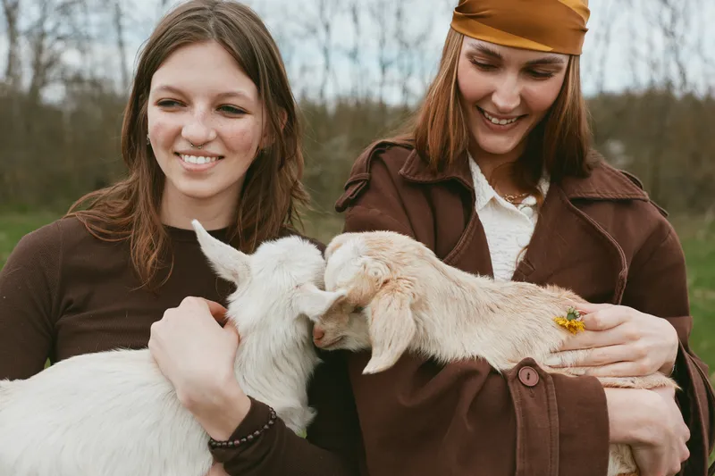Two women outdoors hug white goat kids, sharing gentle affection and smiles, wearing casual jackets, in a natural park setting, a wholesome lifestyle moment with friendly animals today.