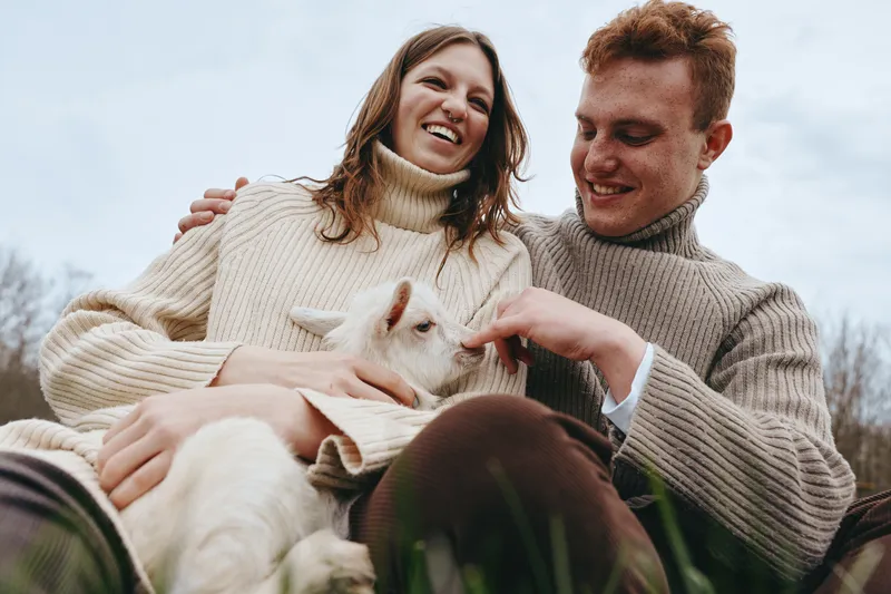 Cheerful couple outdoors, smiling with a gentle goat kid between them, cozy sweaters, sunlit moment, soft breeze, natural colors, warm connection, rural backdrop, candid lifestyle scene