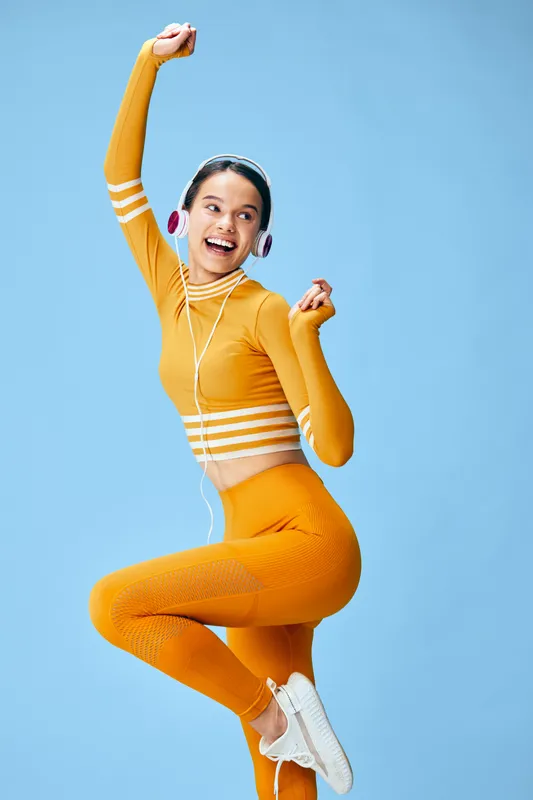 Young woman fitness and sport model in vibrant yellow sportswear jumps mid-air with white sneakers and headphones, smiling energetically against a clean blue studio background conveying vitality.