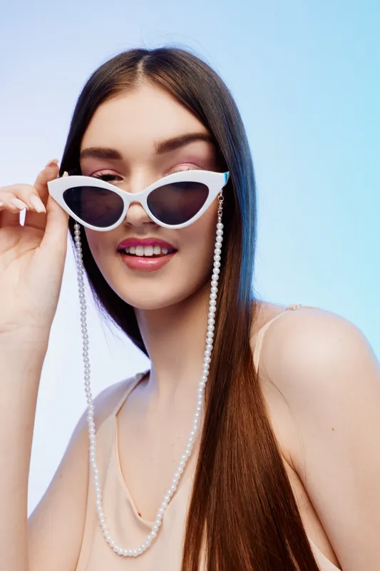 Young woman with long hair wearing white cat eye sunglasses and a pearl chain smiles confidently on a blue background. Concept of beauty fashion and stylish accessories.