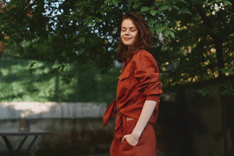 Young woman with wavy brown hair stands outdoors in a red outfit, smiling softly as she enjoys a sunlit moment among green trees