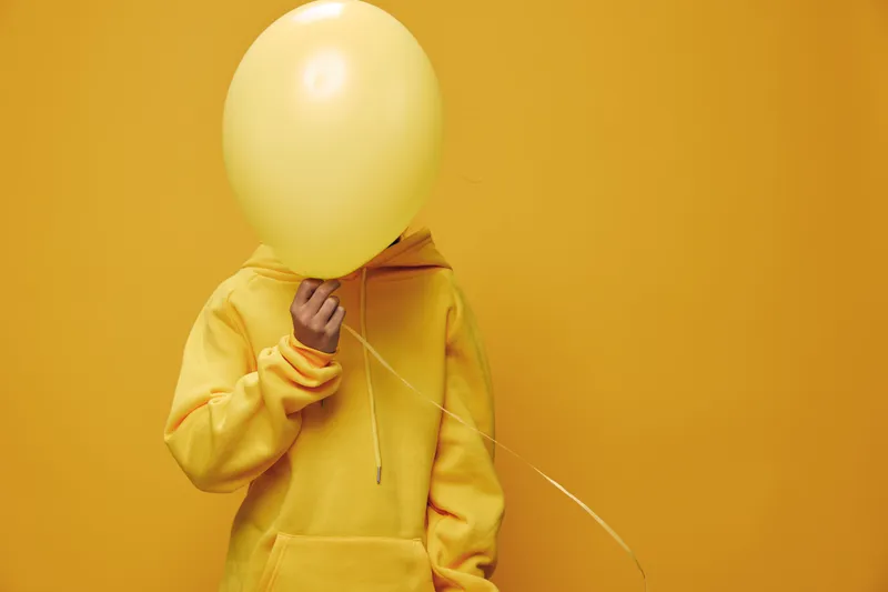 Person in yellow hoodie holding yellow balloon in front of face with yellow background. Monochrome style, casual clothing, isolated portrait for concept or creativity.