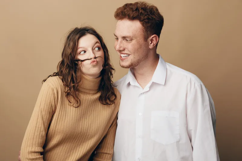 Playful young woman with curly hair makes funny face using hair while smiling man in white shirt looks at her. Casual studio portrait on beige background. Concept of friendship and humor.