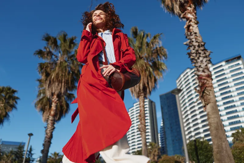 A woman in a red coat walks outdoors, gliding through palm trees with modern buildings in the background, capturing a joyful, sunlit city moment