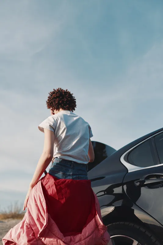 Woman in a flowing dress stands beside a sleek car in bright outdoor light back view fashion moment urban road scene sense of freedom and movement