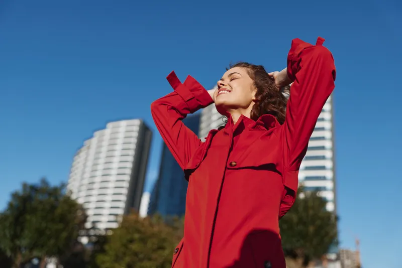 Happy woman in red jacket enjoying sunny day outdoors with eyes closed and hands behind head. Urban background with blue sky and modern buildings.