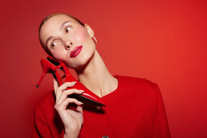 Stylish woman in red sweater holds red high heel shoe near face, posing against red background. Concept of fashion, style, elegance, and bold color contrast in studio lighting.