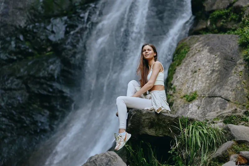 woman sitting by waterfall in white activewear with sneakers, serene nature background, outdoor relaxation and fitness lifestyle scene