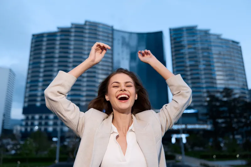 Happy woman celebrating success outdoor in urban business district with modern high-rise buildings, joyful expression, beige jacket, evening lighting