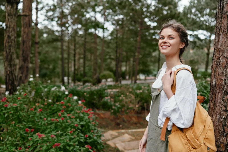 Young woman enjoying nature in a green park, wearing a bright yellow backpack, smiling, surrounded by vibrant flowers and tall trees, reflecting a joyful atmosphere