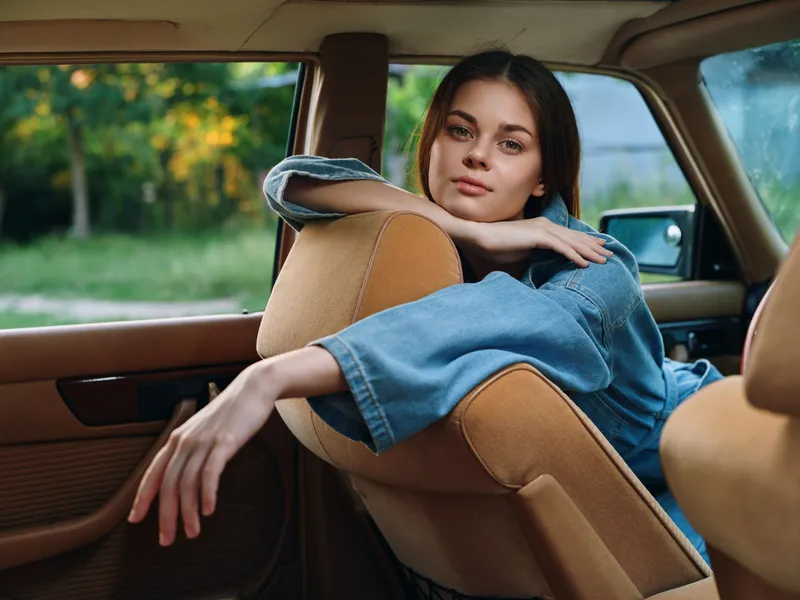 Young woman in denim jacket sitting in a vintage car, relaxed and contemplative, with soft natural light and a green outdoor background, evoking a sense of nostalgia.