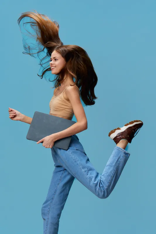 young woman running energetically holding laptop, with flowing hair and casual denim outfit against blue studio background, conveying dynamic motion and joy