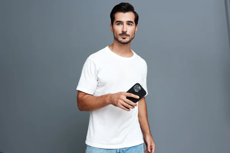 Smiling young man in white t-shirt holds smartphone, looking at the camera with a cheerful expression, captured against a grey background. Lifestyle concept.
