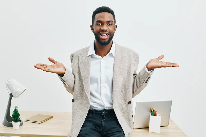 Confused man with open arms in office environment, wearing smart casual blazer and white shirt, standing at desk with laptop and stationery, expressing uncertainty or questioning, indoor workspace, pr