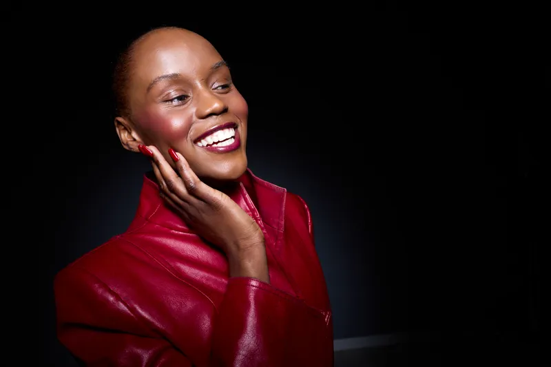 Smiling black woman with dark skin wearing red leather jacket touches face with manicured hand. Studio portrait on black background. Concept of beauty, fashion, confidence, and self-care.