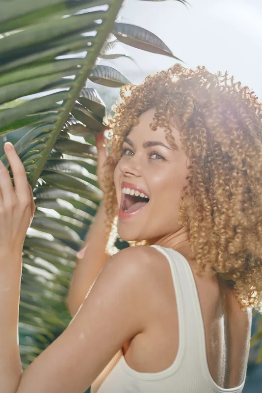 happy woman with curly hair smiling brightly near tropical leaves in bright natural light, summertime outdoor lifestyle, joyful expression
