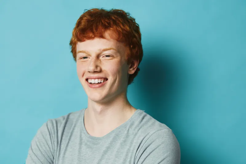 Redhead man with curly hair wearing gray t shirt smiling and looking away on blue background. Male portrait with casual style and natural expression for happiness and youth.