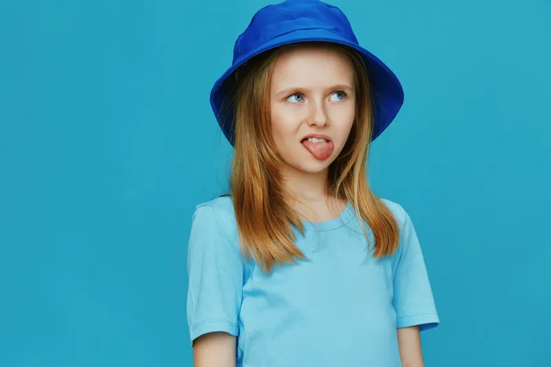 Playful young girl with long hair wearing a blue bucket hat and t shirt, expressing a fun emotion against a vibrant blue background in a colorful and cheerful setting