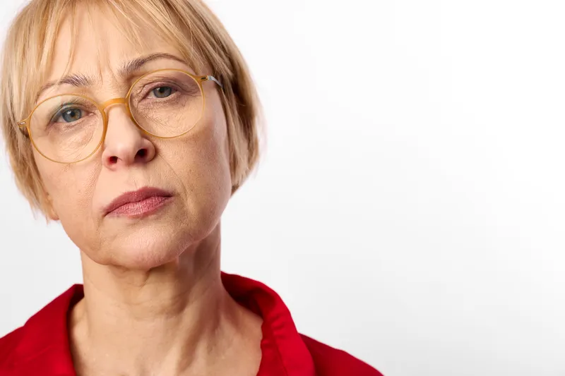 Senior woman portrait, amber glasses on face, neutral thoughtful expression, closeup studio shot, white background, red collar top, healthcare, vision