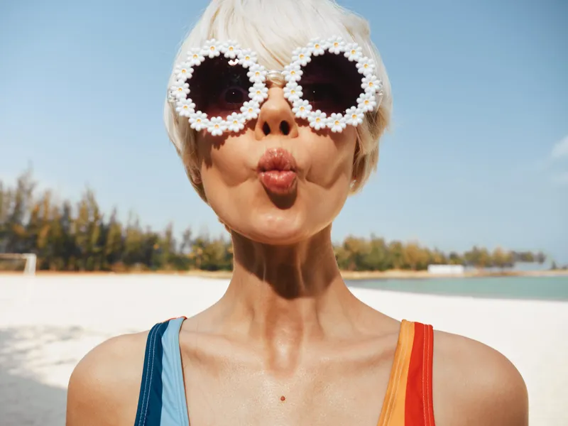 Sunny portrait of a woman at the beach wearing playful flower sunglasses, lips puckered in a kiss, bright swimsuit straps glow under warm sunlight and sea breeze.