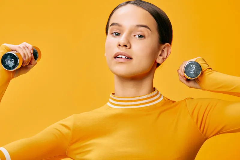 Woman fitness enthusiast lifts dumbbells in a bright orange studio, showcasing strength, athletic training, and a vibrant workout scene with focused expression.