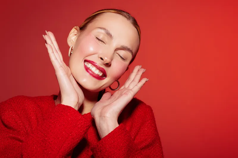 Smiling woman with closed eyes wearing red sweater and lipstick poses happily with hands near face on red background. Concept of joy, beauty, fashion, and positive emotions.