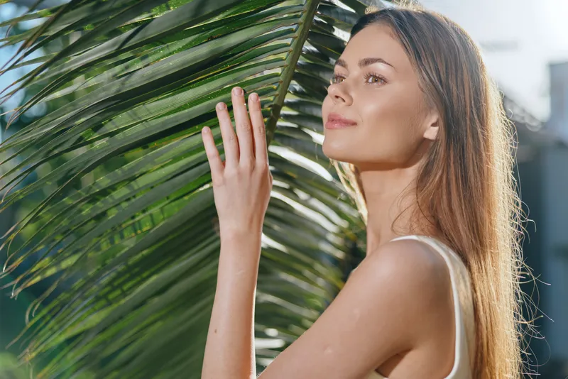 Young woman touching green palm leaves outdoors with soft sunlight, natural beauty, serene expression, summer mood, fresh tropical environment.