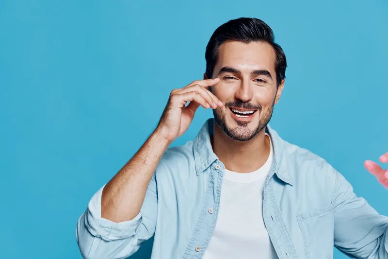 Smiling man wearing a light blue shirt, showcasing a cheerful expression while gesturing playfully against a bright blue background. Portrait with positive vibes.
