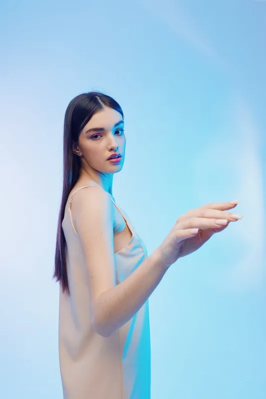 Young woman with long dark hair wearing a beige dress poses gracefully under blue lighting. Concept of beauty, fashion, and elegance in a studio setting.