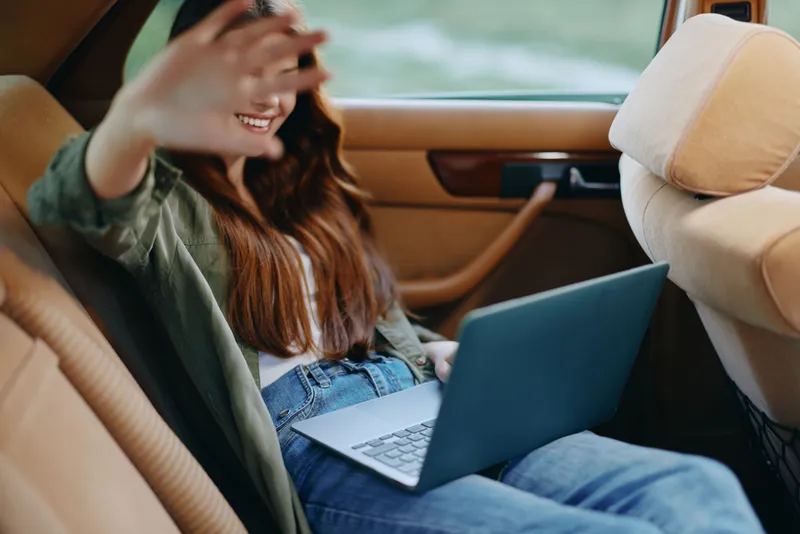 Young woman smiling and working on a laptop in a car, casual attire, natural background, relaxed vibe, showcasing remote work lifestyle.