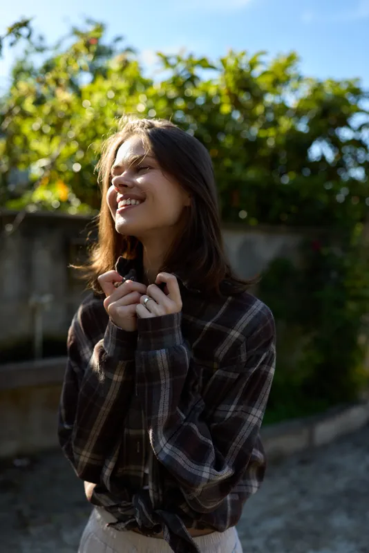 Joyful woman outdoors, smiling softly in sunlight, relaxed pose, plaid shirt, natural colors, lush greenery background, candid lifestyle moment