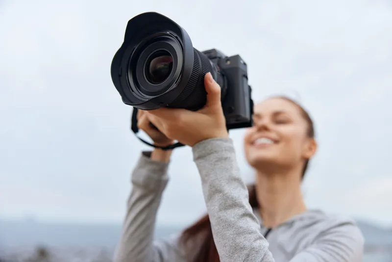Smiling woman holding a camera outdoors, showcasing passion for photography in a casual outfit against a cloudy sky background