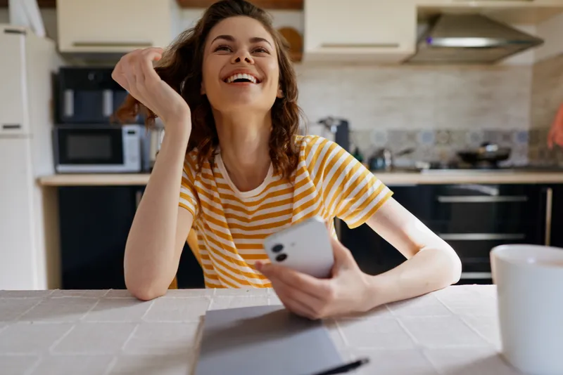 Vibrant woman indoors in a bright kitchen, smiling while looking at her phone, casual mood, home lifestyle moment, sunlit atmosphere, natural colors, cozy setting
