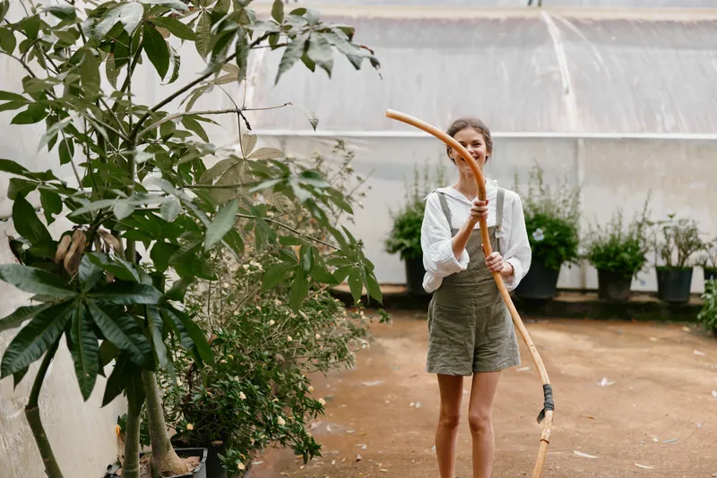 Young woman in light clothing smiling while holding a long garden tool in a greenhouse surrounded by lush green plants and soft natural light
